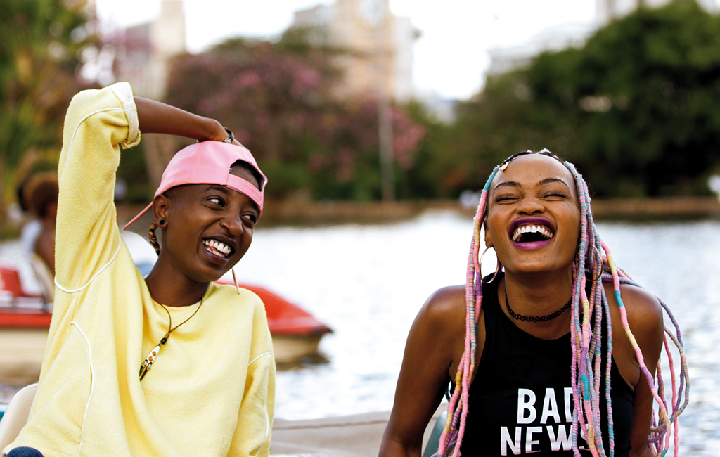 Image is from the film 'Rafiki' (2018). Two women laugh whilst peddling a boat on a lake. One is wearing a yellow jumper and a pink shirt whilst the other has pastel coloured dreadlocks and is wearing a black vest top.